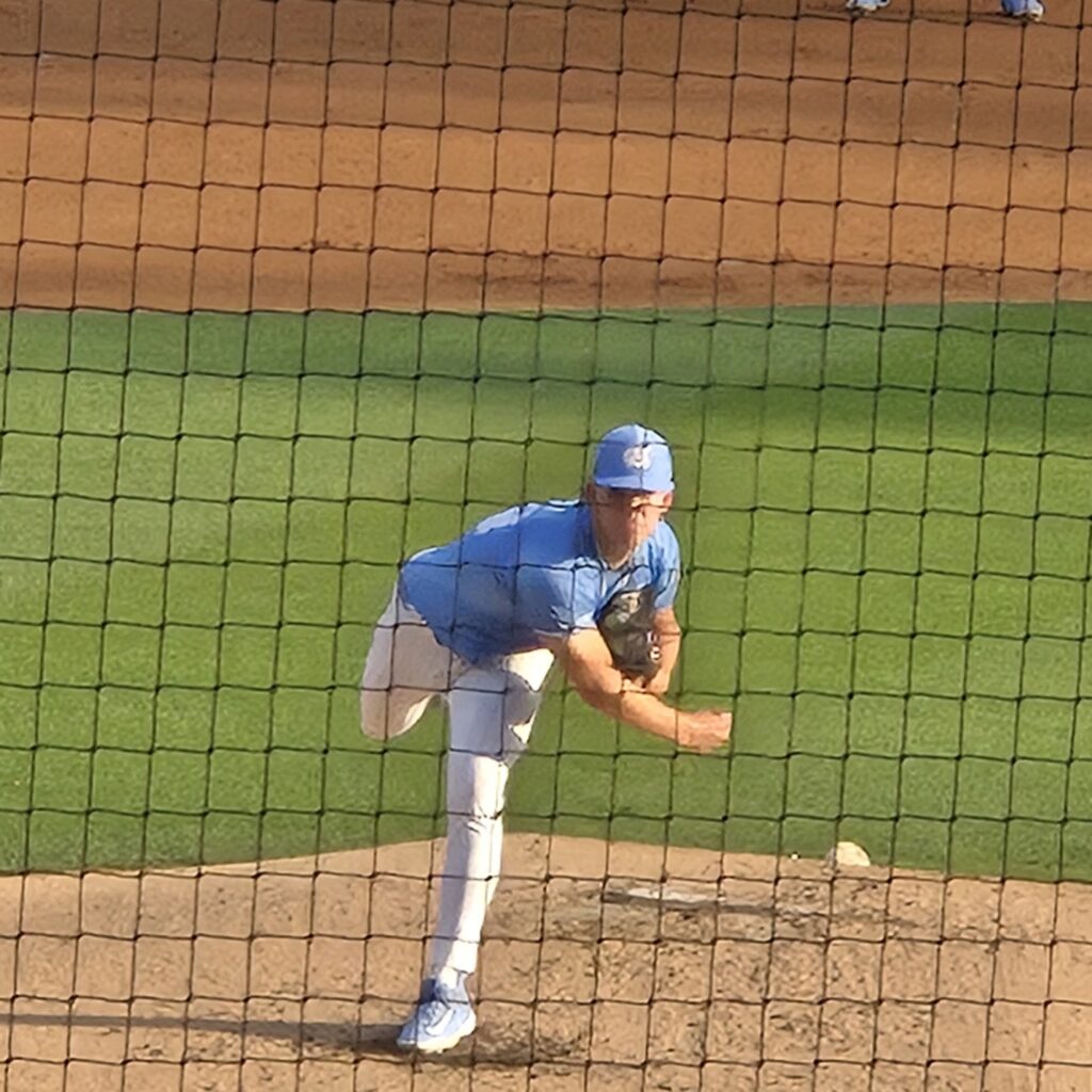 Baseball pitcher in light blue uniform delivering a pitch from the mound, behind home plate net.