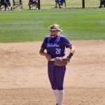 Baseball player in purple pinstripe uniform no. 26, wearing a catcher's mask and glove, standing on the infield dirt.