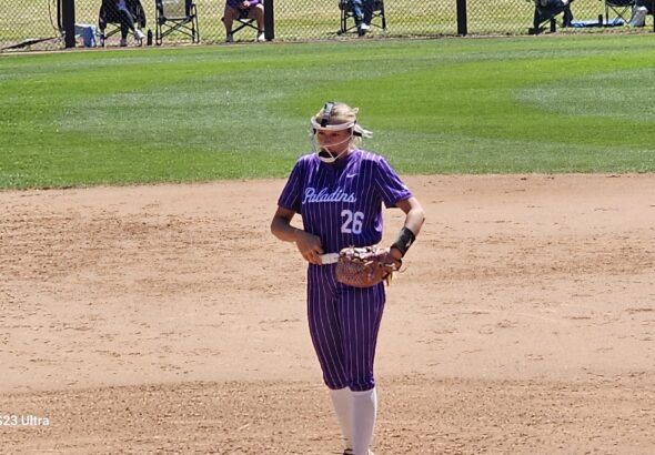 Baseball player in purple pinstripe uniform no. 26, wearing a catcher's mask and glove, standing on the infield dirt.