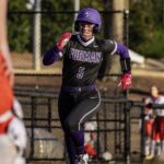 Furman softball player in uniform #5 sprinting with a ball in hand during a game, wearing a purple helmet and black–purple gear.