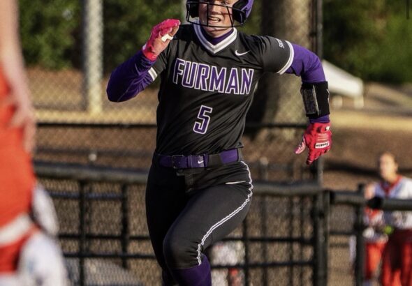 Furman softball player in uniform #5 sprinting with a ball in hand during a game, wearing a purple helmet and black–purple gear.