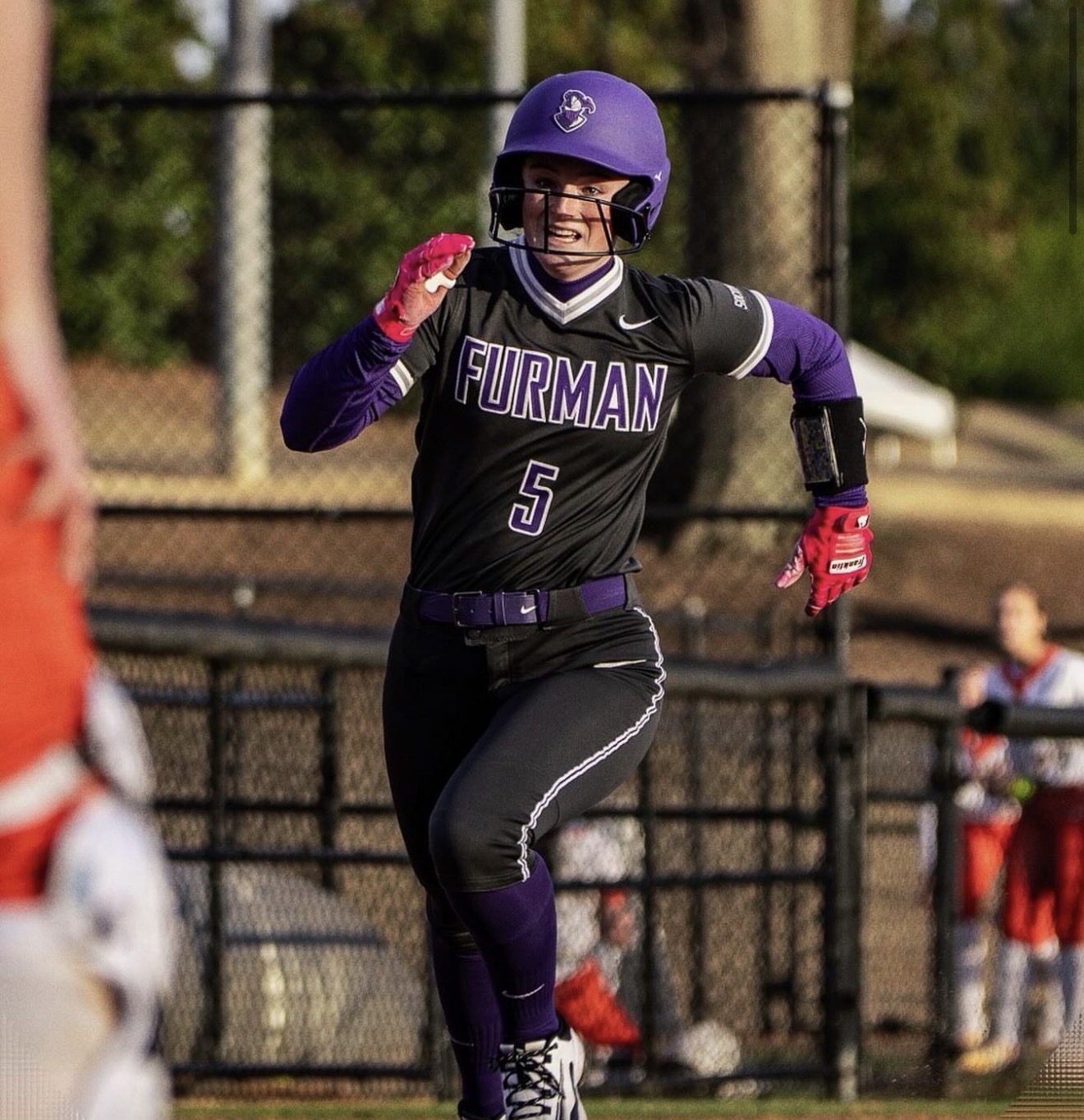 Furman softball player in uniform #5 sprinting with a ball in hand during a game, wearing a purple helmet and black–purple gear.