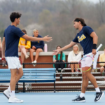Two male tennis players on a blue court greet with a high-five after a match, spectators watching from the bleachers in the background.
