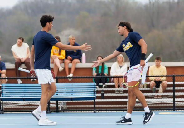Two male tennis players on a blue court greet with a high-five after a match, spectators watching from the bleachers in the background.