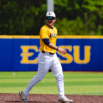 Baseball player in a yellow BUCS jersey and white pinstriped pants walks on the infield dirt, wearing a cap and sunglasses, with a blue outfield wall behind.