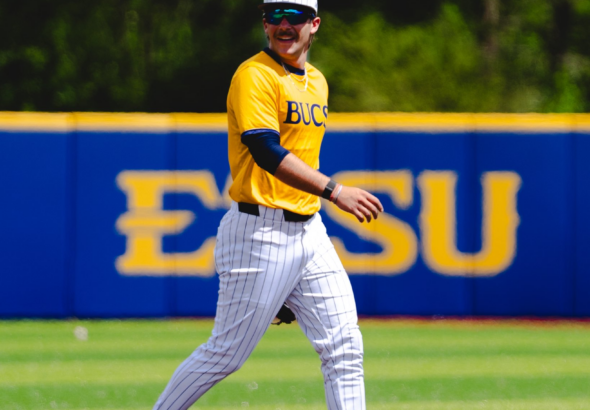 Baseball player in a yellow BUCS jersey and white pinstriped pants walks on the infield dirt, wearing a cap and sunglasses, with a blue outfield wall behind.