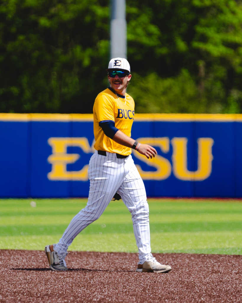 Baseball player in a yellow BUCS jersey and white pinstriped pants walks on the infield dirt, wearing a cap and sunglasses, with a blue outfield wall behind.