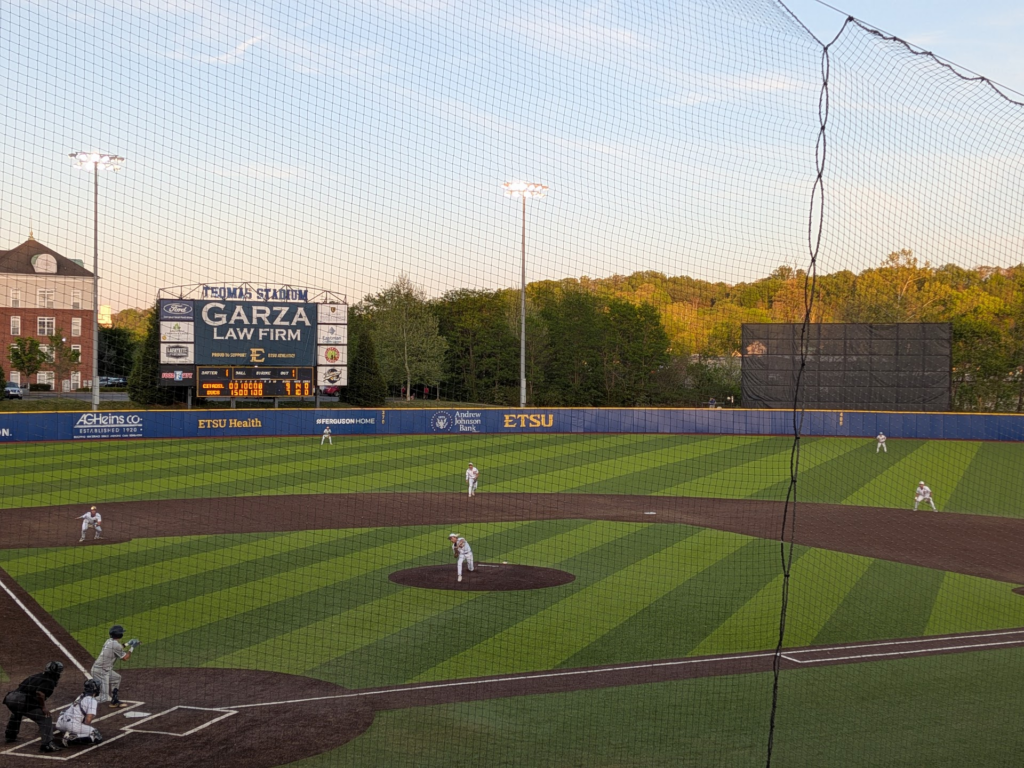 Baseball game at Thomas Stadium; scoreboard reads Garza Law Firm with ETSU banners along the outfield wall and players on the fieldpl