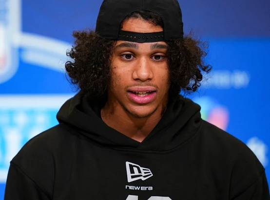 Young male athlete in a black hoodie and backward cap speaks at a press conference; NFL microphone in front.
