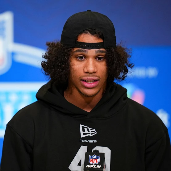 Young male athlete in a black hoodie and backward cap speaks at a press conference; NFL microphone in front.