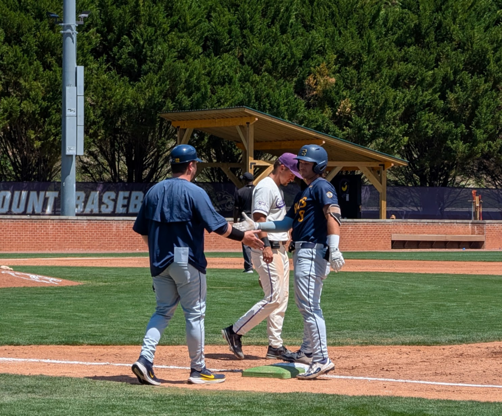 Baseball players greet with a high-five near first base on a sunny field, with a coach watching in blue.