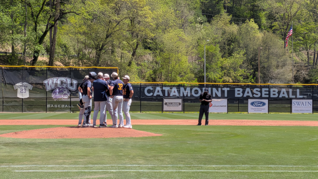Baseball players huddle on the mound with banners and an American flag in the outfield background.