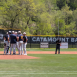 Baseball players huddle on the mound with banners and an American flag in the outfield background.
