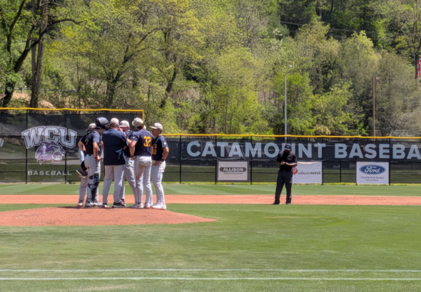 Baseball players huddle on the mound with banners and an American flag in the outfield background.