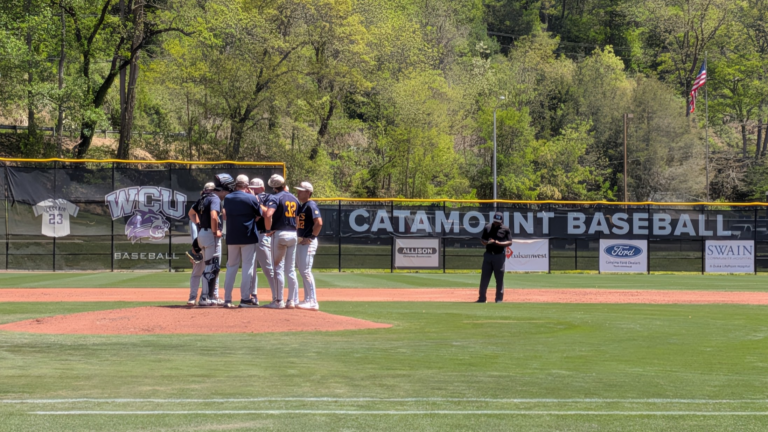 Baseball players huddle on the mound with banners and an American flag in the outfield background.