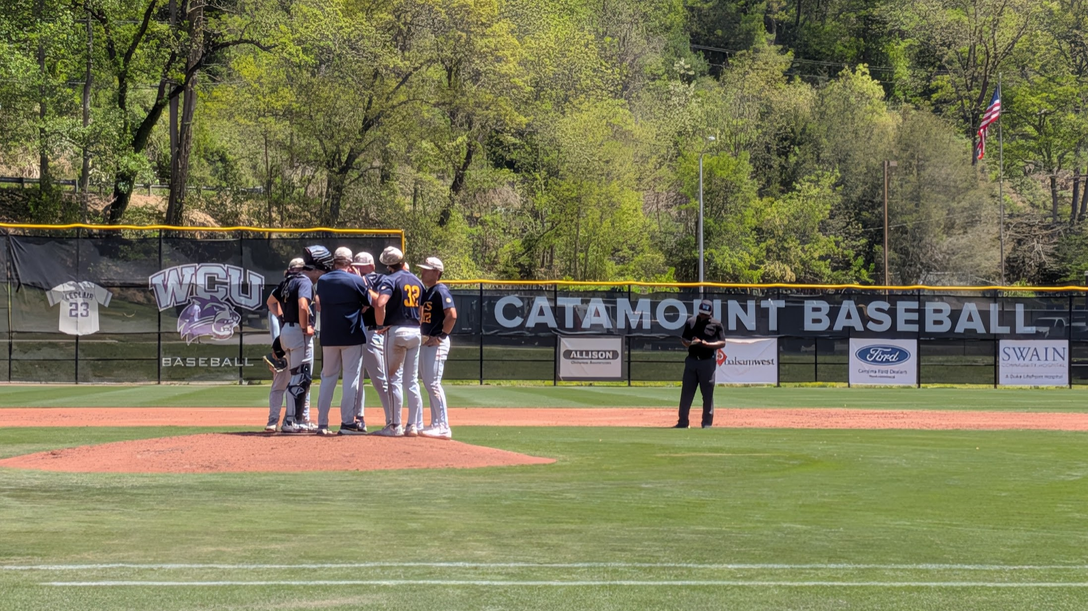 Baseball players huddle on the mound with banners and an American flag in the outfield background.