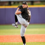 Baseball pitcher in black uniform on the mound mid-pitch, wearing a WF cap and gray pants against a purple outfield wall.