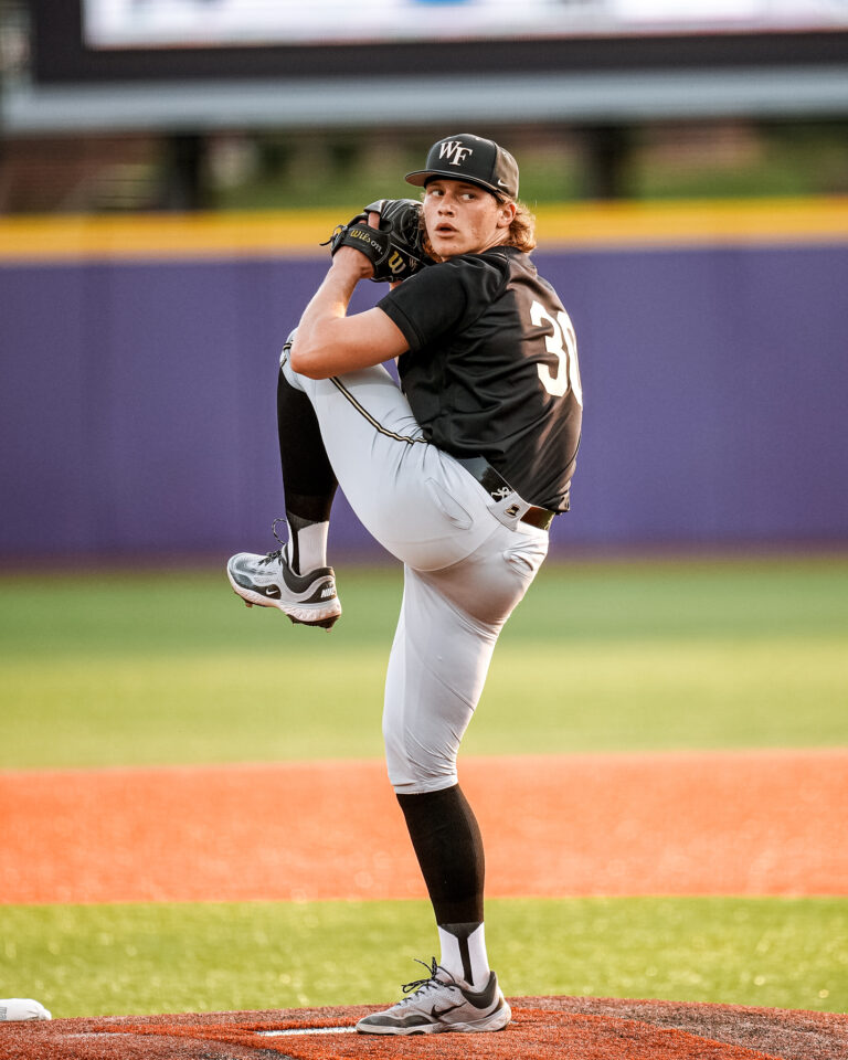 Baseball pitcher in black uniform on the mound mid-pitch, wearing a WF cap and gray pants against a purple outfield wall.