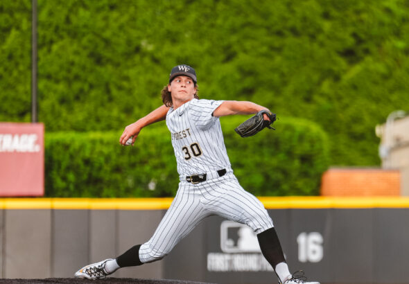 Baseball pitcher in a white pinstripe uniform throwing from the mound with a black cap and glove, mid-action against a green outfield backdrop.