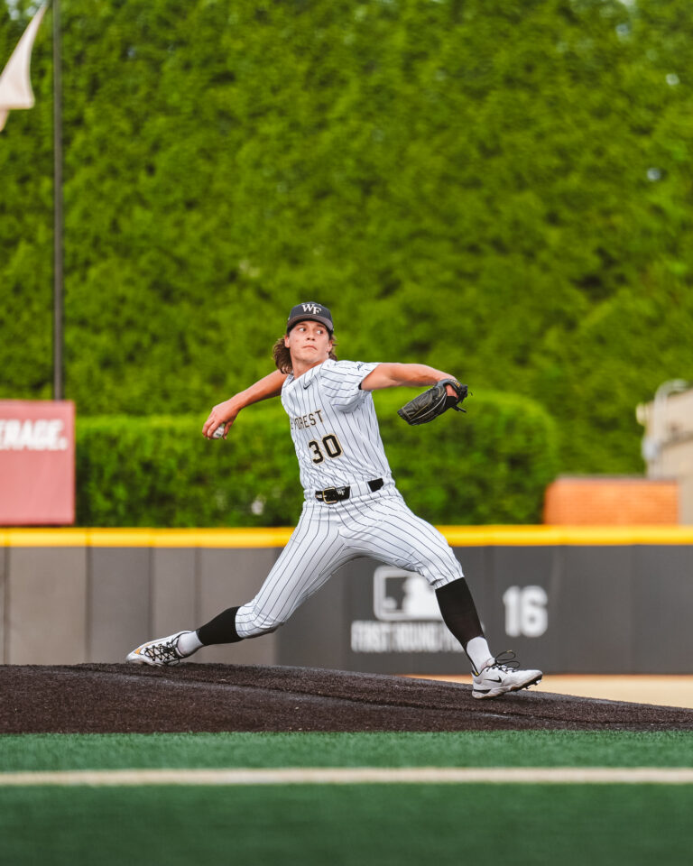 Baseball pitcher in a white pinstripe uniform throwing from the mound with a black cap and glove, mid-action against a green outfield backdrop.
