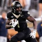 African American football player in a black uniform running with the ball on a football field.