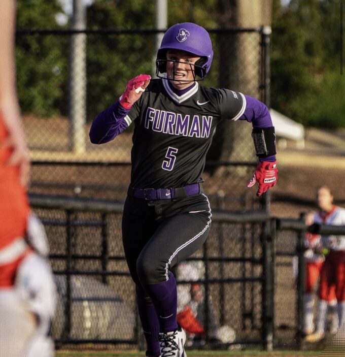 Furman softball player in a black jersey with number 5, purple helmet and gloves, running on the field.