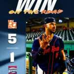 Baseball player in a blue jersey and cap walks out from the dugout on a promotional poster that reads 'WIN ON THE ROAD' at the top.
