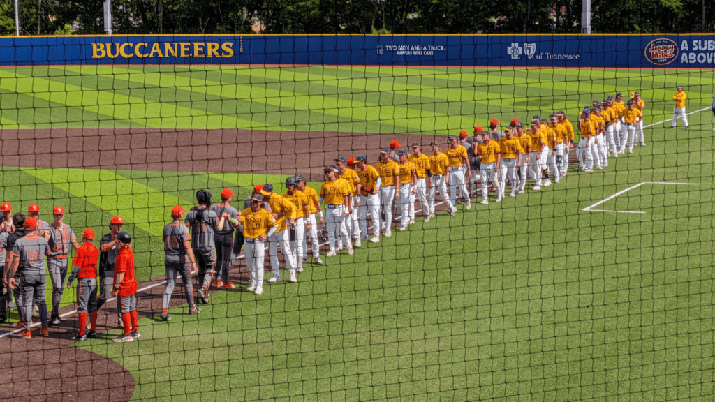 Baseball players in yellow jerseys form a line along the first-base line as a game is about to start, behind a blue outfield wall labeled Buccaneers.