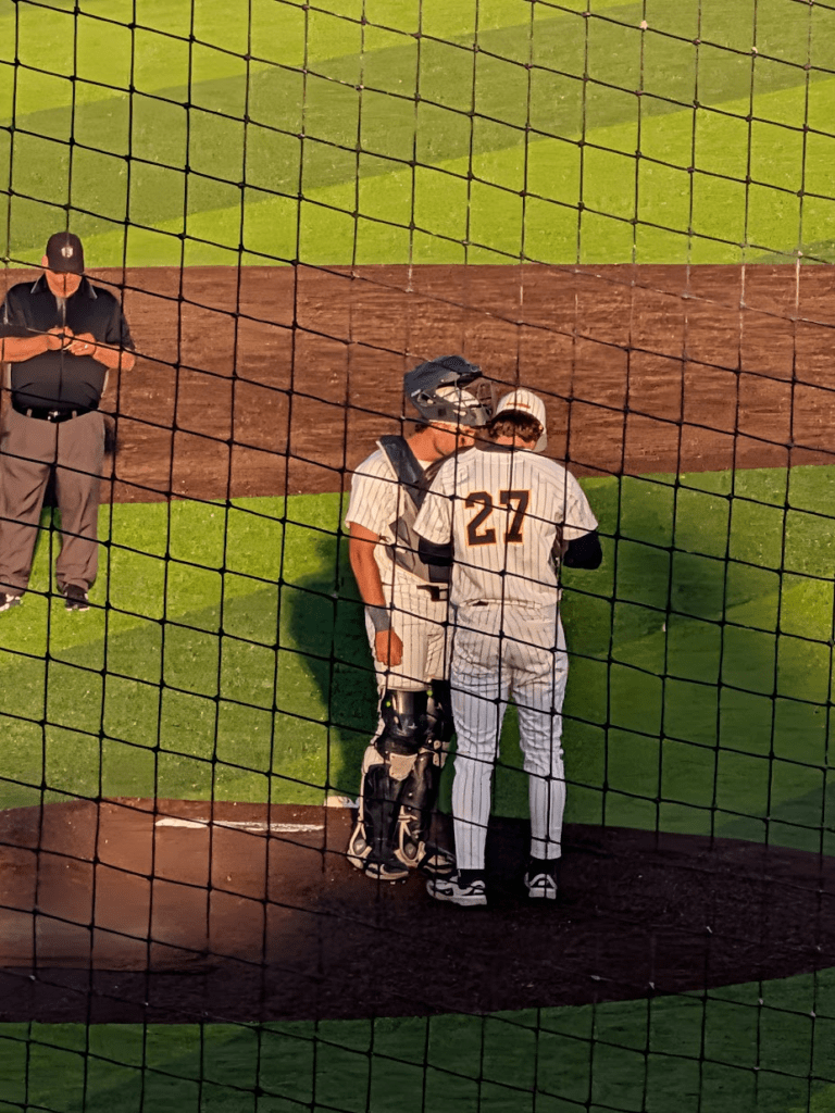 Two baseball players at home plate; batter wearing #27 and catcher in full gear, talking as an umpire watches behind the netting.