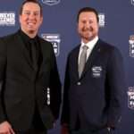 Two men in formal suits posing on a blue NASCAR Hall of Fame backdrop at the Forever Legends event.