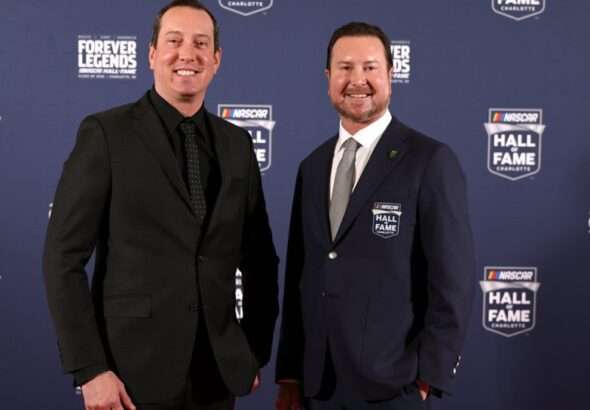 Two men in formal suits posing on a blue NASCAR Hall of Fame backdrop at the Forever Legends event.