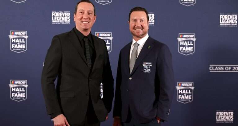 Two men in formal suits posing on a blue NASCAR Hall of Fame backdrop at the Forever Legends event.