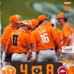 Group of college baseball players in orange uniforms huddle on the field; score shows 4-8 with team logos at bottom. The back of player Clark #16 is visible.