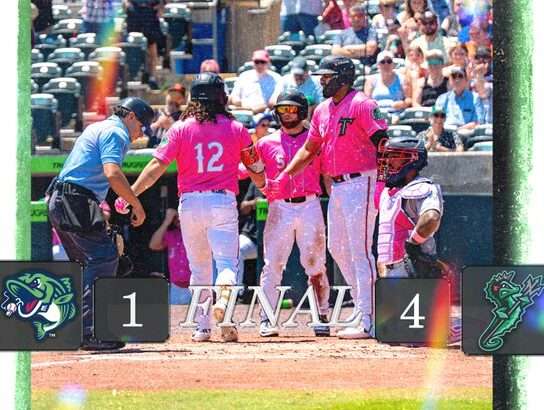 Baseball players in pink uniforms celebrate on the field after a 4–1 victory; final score displays 1–4 with team logos visible.