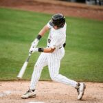 Baseball player in striped uniform swings at a pitched ball near home plate on a green field.