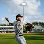Baseball pitcher in mid-pitch on a sunny field wearing a WAKE uniform, ball in hand ready to throw.