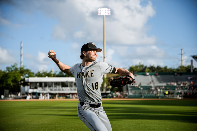 Baseball pitcher in mid-pitch on a sunny field wearing a WAKE uniform, ball in hand ready to throw.