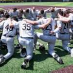 Wake Forest baseball players in gray pinstripe uniforms form a circle huddle on the field, arms around each other.
