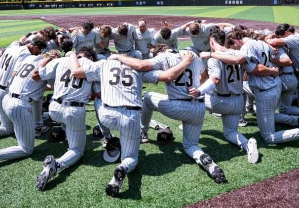 Wake Forest baseball players in gray pinstripe uniforms form a circle huddle on the field, arms around each other.