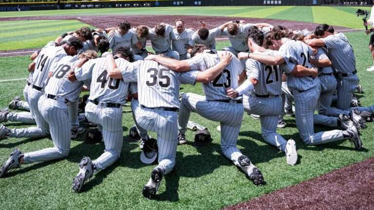 Wake Forest baseball players in gray pinstripe uniforms form a circle huddle on the field, arms around each other.