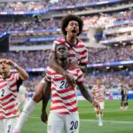 USA soccer players celebrate a goal on the field, one player carried on another's shoulders as fans cheer in the stands ahead of them on the green pitch.