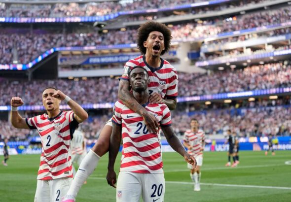 USA soccer players celebrate a goal on the field, one player carried on another's shoulders as fans cheer in the stands ahead of them on the green pitch.