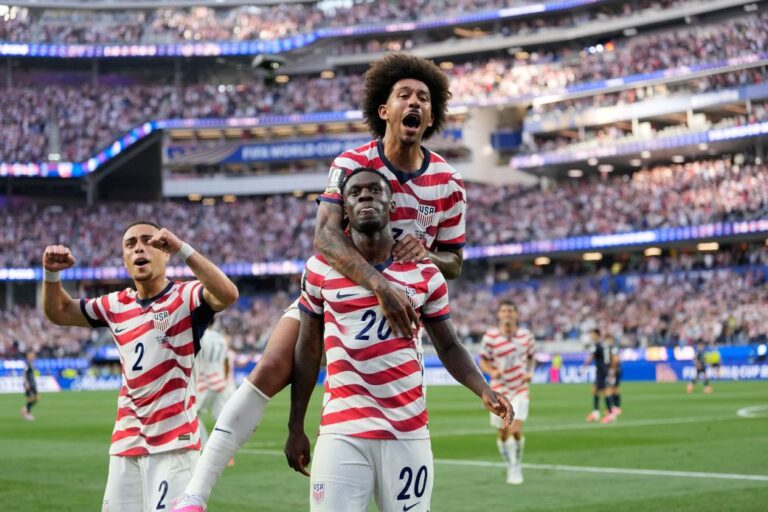 USA soccer players celebrate a goal on the field, one player carried on another's shoulders as fans cheer in the stands ahead of them on the green pitch.