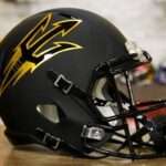 Black football helmet with a yellow stylized bolt/logo on a wooden table surface, sans-face shield in foreground.