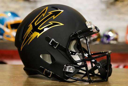 Black football helmet with a yellow stylized bolt/logo on a wooden table surface, sans-face shield in foreground.
