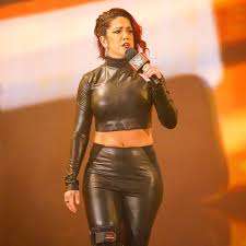 Female singer in a black leather two-piece outfit on stage, holding a microphone under orange stage lights.