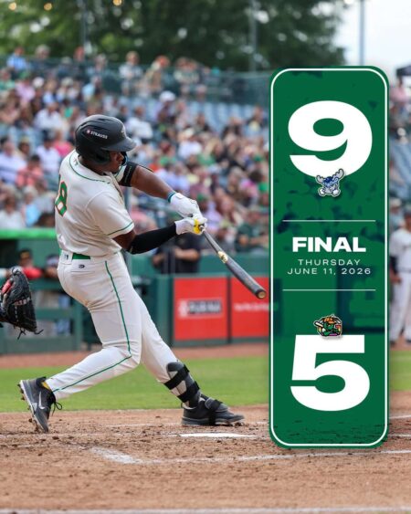 Batter in a white baseball uniform swings at a pitch in the batter's box with crowds in the stands; a tall green final banner stands to the right with a large 9 and 5 and the date June 11, 2026.