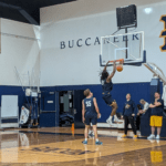 Player in a blue uniform dunks the ball during practice in a gym with 'BUCCANEER' lettering on the wall.