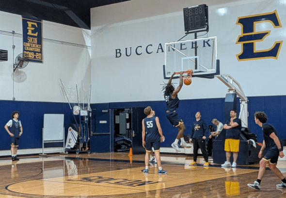 Player in a blue uniform dunks the ball during practice in a gym with 'BUCCANEER' lettering on the wall.