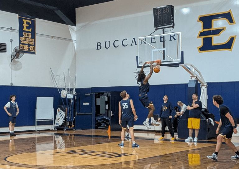 Player in a blue uniform dunks the ball during practice in a gym with 'BUCCANEER' lettering on the wall.